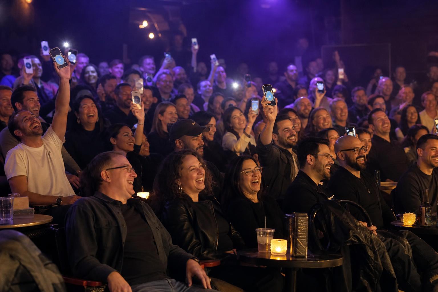 Laughing audience holding up glowing phones to vote during a live comedy show