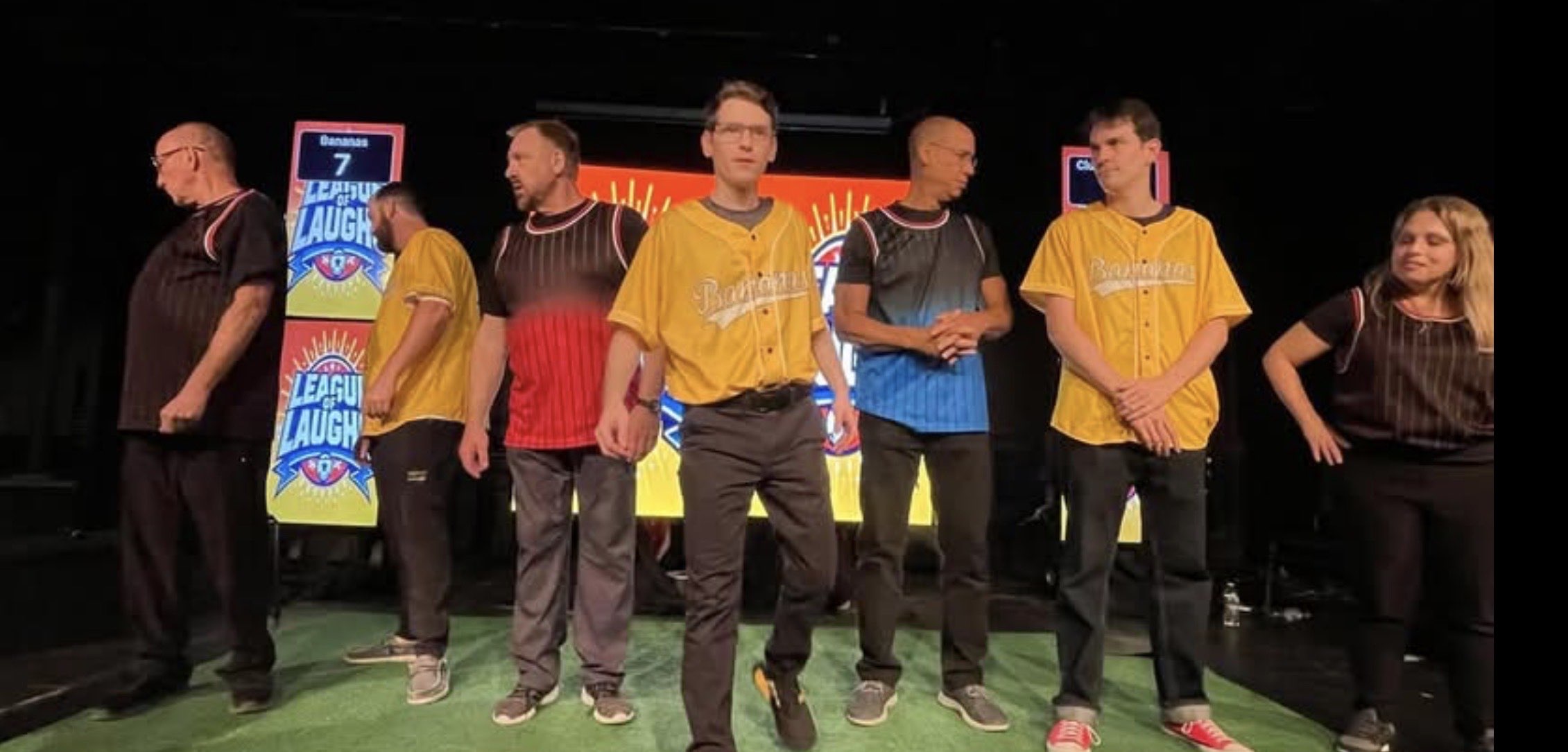 Seven Comedy Loco performers on a League of Laughs stage: yellow Bananas jerseys versus dark jerseys, with League of Laughs banners and a Bananas 7 scoreboard